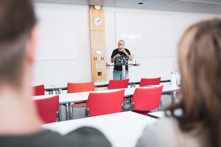 Florian speaking at a podium in a lecture hall at the University of Bayreuth, gesturing next to a laptop covered in stickers, with a terminal-styled slide reading "Fragen?" projected behind him and listeners in the foreground.