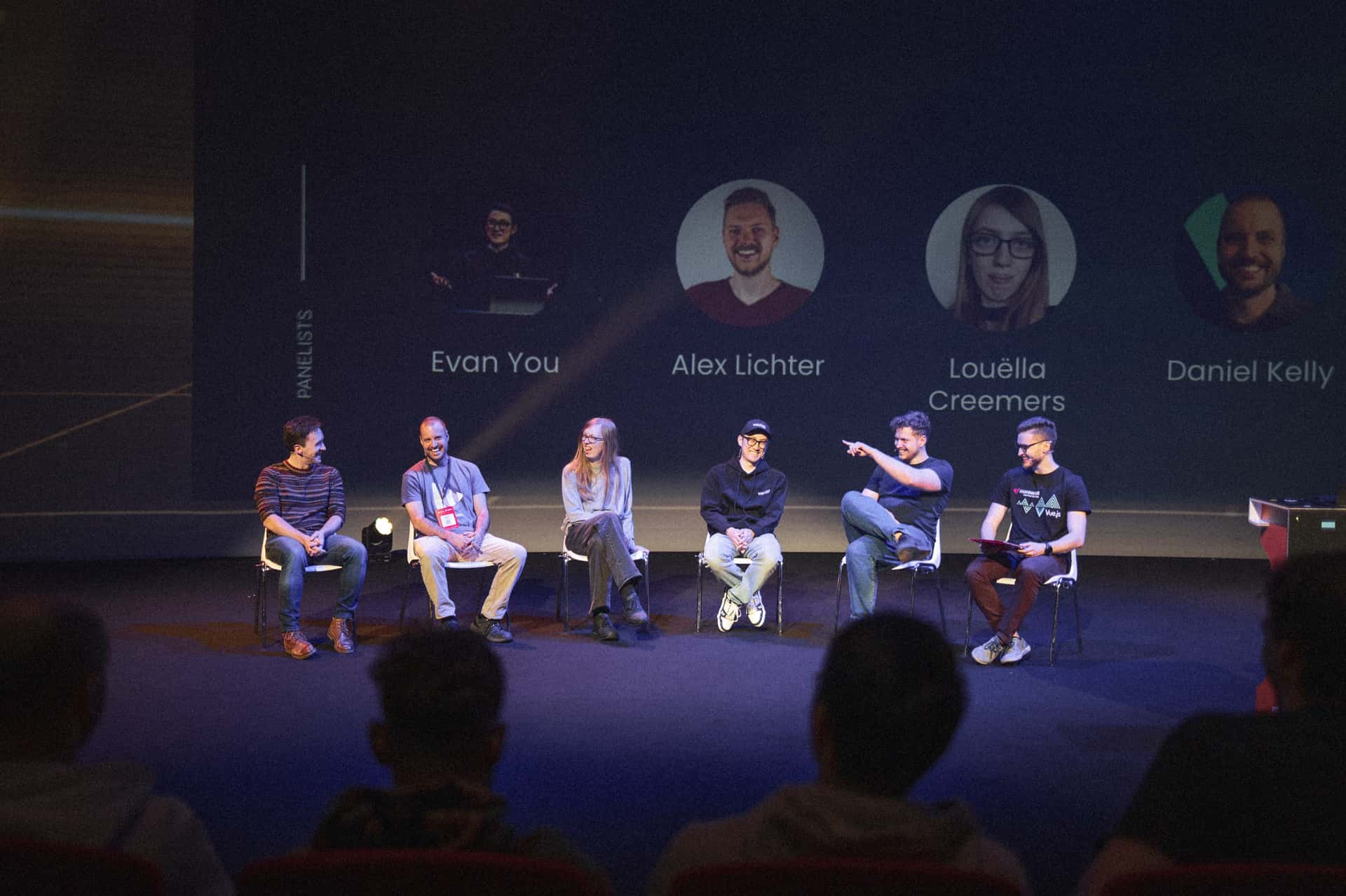 A panel of six individuals (Daniel Roe, Daniel Kelly, Louëlla Creemers, Evan You, Alexander Lichter and Jakub Andrzejewski) is seated on stage at Vue.js Amsterdam, engaging in discussion during the panel discussion. Each panelist appears to be sharing insights, with two of them actively gesturing. The background features names and images of the panelists displayed prominently, enhancing the setting. The audience is partially visible, seated in the foreground, indicating an interactive environment. The lighting is focused on the panel, creating an inviting atmosphere for conversation.